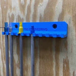 以下が含まれることがあります： 「FGS ENGINEERING」という文字が書かれた青い3Dプリントの壁掛けに、金属製の棒と黄色の工具がいくつか取り付けられています。