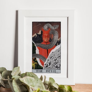 May include: A framed collage art print of a tarot card, The Devil, featuring a red figure with a snake wrapped around its head. The background is a collage of different textures and colors.