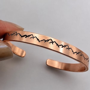 May include: Copper cuff bracelet featuring a mountain range design. The open cuff is a warm copper color with a black mountain silhouette etched along the top. The bracelet is held by a hand.