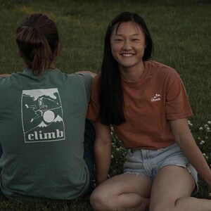 May include: Two people sitting on grass, one wearing a green t-shirt with a white graphic and the word "CLIMB". The other person is wearing an orange t-shirt with a small white logo. Both are smiling.