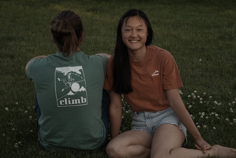 May include: Two people sitting on grass, one wearing a green t-shirt with a white graphic and the word "CLIMB". The other person is wearing an orange t-shirt with a small white logo. Both are smiling.