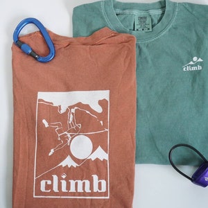 May include: Two t-shirts, one rust-colored and one teal, with climbing-themed graphics. The rust shirt features a climber graphic and the word "climb." A blue carabiner and a purple climbing accessory are also present.