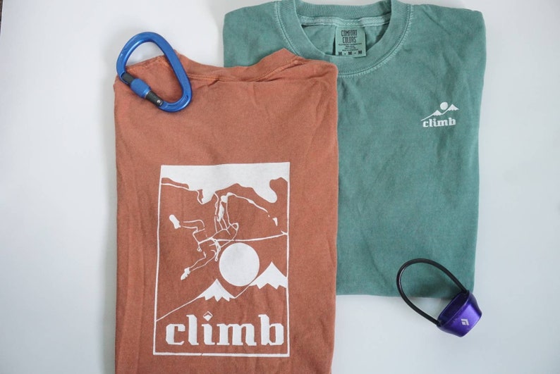 May include: Two t-shirts, one rust-colored and one teal, with climbing-themed graphics. The rust shirt features a climber graphic and the word "climb." A blue carabiner and a purple climbing accessory are also present.