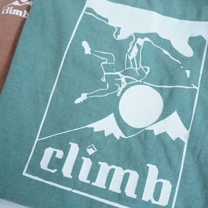 May include: Two t-shirts, one in a muted rose color and the other in a teal green, are displayed. The green shirt features a white graphic of a climber, mountains, and the word "climb".
