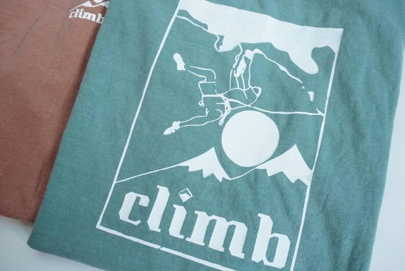May include: Two t-shirts, one in a muted rose color and the other in a teal green, are displayed. The green shirt features a white graphic of a climber, mountains, and the word "climb".