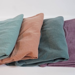 May include: Four folded sweatshirts in shades of teal, peach, dark teal, and purple. The sweatshirts are made of a soft material and are neatly stacked against a white background. The image highlights the variety of colors available.
