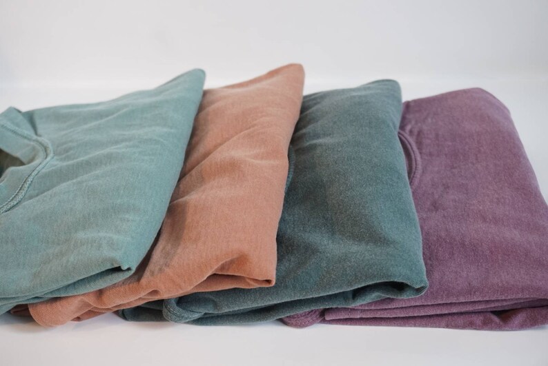 May include: Four folded sweatshirts in shades of teal, peach, dark teal, and purple. The sweatshirts are made of a soft material and are neatly stacked against a white background. The image highlights the variety of colors available.