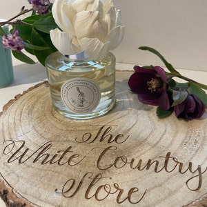 May include: A white ceramic flower diffuser with a clear glass bottle filled with liquid. The bottle has a label that reads "The White Country Store" and "English Plum". The diffuser is sitting on a wooden slice with the words "The White Country Store" engraved on it.