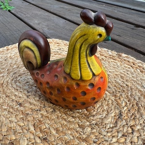 May include: A carved wooden rooster figurine featuring a yellow neck, a dark brown comb and tail, and an orange body with circular indentations. The surface displays a textured finish with visible wood grain and hand-painted color gradients.