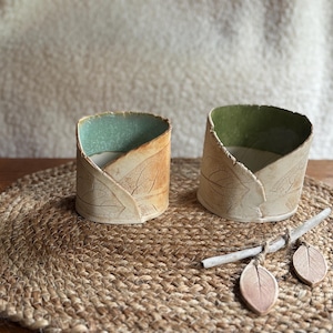 May include: Two ceramic leaf-textured candle holders with teal and green interiors sit on a woven mat. A small decorative twig with two leaf-shaped ornaments rests nearby. The candle holders have a natural, earthy aesthetic.