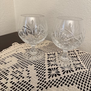 May include: Two clear crystal brandy snifters with intricate starburst patterns on the bowl. The glasses sit on a cream-colored, crocheted doily. The stemware is designed for serving brandy or other spirits.