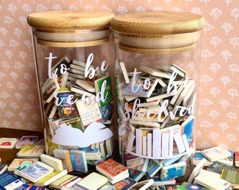 May include: Two glass jars with wooden lids filled with miniature book covers. The jars have white vinyl lettering that reads "to be read" and "to be shelved".