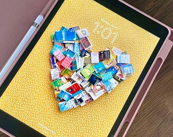 Pode incluir: Um tablet digital com fundo amarelo e um arranjo em forma de coração de livros em miniatura. A tela do tablet mostra a data "Segunda-feira, 17 de abril" e a hora "1:01".