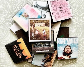 May include: A collection of vintage music album covers featuring various artists and genres. The covers are in different colors and styles, including a black and white photo of a band, a colorful illustration of a cartoon character, and a photo of a group of people in a field. The album covers are arranged in a pile on a patterned surface.
