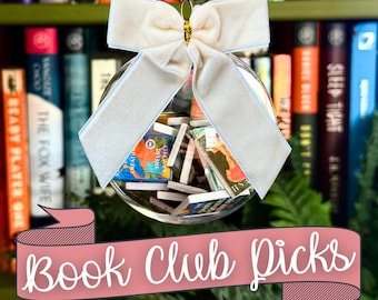 May include: A clear spherical ornament filled with miniature books, topped with a light cream-colored velvet bow. The ornament hangs by a silver cord. A pink banner with the white text "Book Club Picks" is overlaid at the bottom of the image.