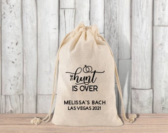 May include: A white cotton drawstring bag with black text that reads "The hunt is over" and "Melissa's Bach Las Vegas 2021".