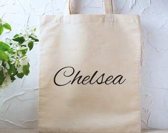 May include: A plain white canvas tote bag with black handles and the name "Chelsea" printed in a cursive font.