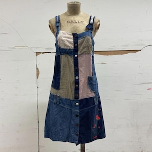 May include: A patchwork denim dress with a button-down front and adjustable straps. The dress features various fabric panels in shades of blue, pink, and tan, with floral embroidery near the hem. The dress is displayed on a mannequin.