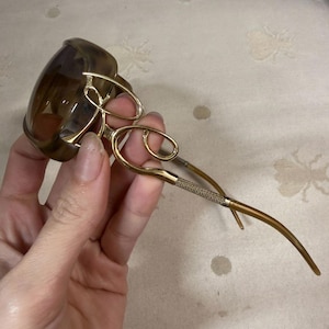 May include: Brown-tinted sunglasses with gold-colored frames. The frames have a decorative, looping design near the lenses and textured detailing on the temples. The lenses are a dark brown color.