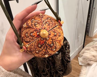 May include: A round, intricately carved wooden purse with a floral design in shades of orange and brown. The purse has a small wooden knob in the center and a dark green strap. The purse is paired with a dark, patterned fabric bag.