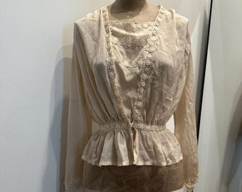 May include: A cream-coloured blouse with long sleeves and a peplum waist. The blouse has delicate lace trim around the neckline and down the front. The garment is displayed on a mannequin.