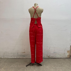 Puede incluir: Un conjunto rojo de dos piezas sobre un maniquí. La parte superior tiene la espalda cruzada y los pantalones son de pierna ancha. La tela tiene un patrón sutil de color más claro. El conjunto se exhibe en un maniquí con torso de color neutro.