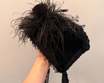May include: A black bonnet with a cluster of dark feathers on top. The bonnet features lace trim and long black ribbons. The hat is a vintage style, likely from the Victorian or Edwardian era.