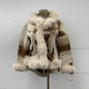 May include: A vintage-style jacket with a cream-colored fur-lined hood, cuffs, and front trim. The jacket's body features a patterned design in shades of brown and cream. The jacket is displayed on a mannequin.
