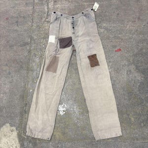 May include: Light gray pants with multiple patches in various shades of brown and white. The pants have a button fly and a tag attached at the top. The pants are made of a soft material.