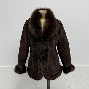 May include: A dark brown suede jacket featuring a thick, plush fur trim along the large shawl collar, the front opening, the sleeve cuffs, and the bottom hem. The jacket has a structured fit with visible seam detailing along the front panels.