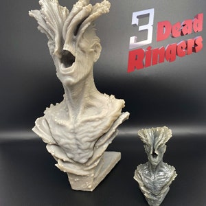 May include: Two 3D printed gray and white busts of a creature with a large mouth and a tree-like head. The larger bust is on a gray base. The smaller bust is on a black surface. The text "3 Dead Ringers" is in the background.