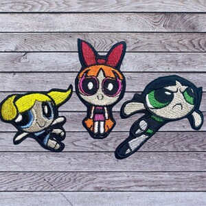 May include: Three embroidered patches featuring characters from The Powerpuff Girls. Bubbles has blonde hair and a blue outfit. Blossom has orange hair and a pink and orange outfit. Buttercup has black hair and a green outfit.