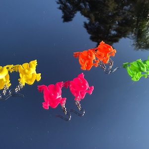 May include: A collection of colorful frog-shaped earrings in yellow, orange, pink, and green. Each pair features a small, detailed frog charm suspended from a silver-tone fish hook. The earrings are displayed on a reflective surface.