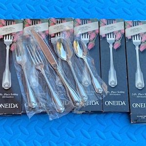 May include: Silverware sets with forks, knives, and spoons are displayed in their original packaging. The boxes are black with the brand name "ONEIDA" and the words "5-Pc. Place Setting". The silverware has a shell design.
