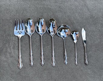 May include: A set of seven silver-plated serving utensils, including a serving fork, a serving spoon, a ladle, a butter knife, and three spoons. The utensils have ornate handles with a floral design.