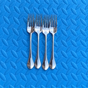 May include: Four polished stainless steel forks arranged in a vertical row. Each fork features a slender handle that widens slightly toward the base and four tines. The metal surface is reflective, showing a smooth, metallic finish throughout.