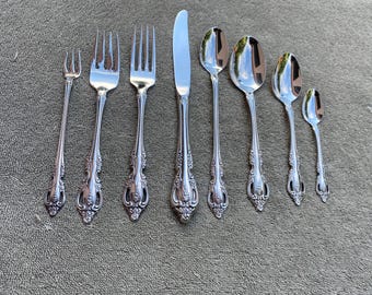 May include: A set of silver-colored flatware, including forks, knives, and spoons, arranged on a textured surface. The utensils have ornate, floral-inspired handle designs. The set includes a variety of spoon sizes, a knife, and several forks.