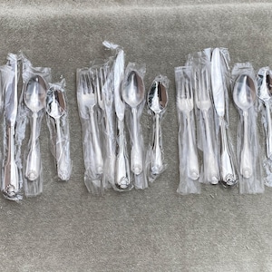 May include: A selection of wrapped cutlery, including forks, spoons, and knives. The silver-coloured utensils are in their original plastic packaging. The arrangement suggests a set of dining cutlery, ready for use.