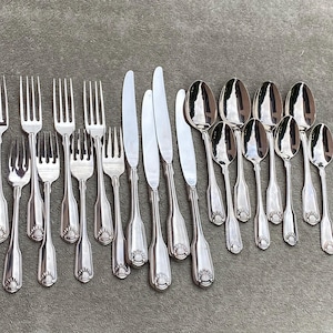 Può includere: Una collezione di posate color argento, tra cui forchette, coltelli e cucchiai, disposte su una superficie grigia. Gli utensili presentano un motivo decorativo alla base dei manici. I cucchiai hanno una superficie riflettente.