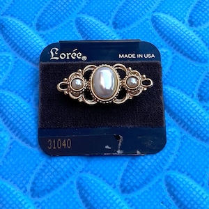 May include: A gold-toned brooch with an oval pearl center and smaller pearl accents. The brooch is on a black velvet backing, with the brand name "Loree" and "MADE IN USA" visible. The item number "31040" is also present.