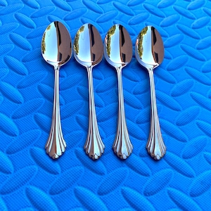May include: Four shiny, silver spoons arranged on a textured blue surface. The spoons have a classic design with a decorative, scalloped pattern at the base of the handle and a rounded bowl. The spoons reflect the surrounding environment.