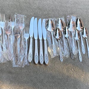 May include: A selection of vintage silverware, including forks, knives, and spoons, displayed on a textured surface. The silver-toned cutlery features decorative handle detailing. Each piece is individually wrapped in clear plastic.