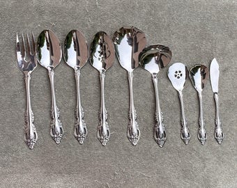 May include: A collection of ornate, silver-colored silverware, including forks, spoons, and serving utensils. The silverware has detailed floral patterns on the handles and is arranged on a neutral-colored surface.