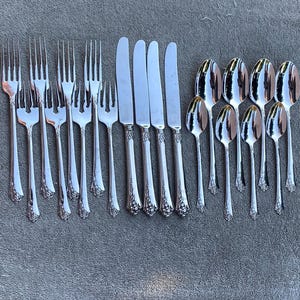 May include: An assortment of silver-coloured cutlery, including forks, knives, and spoons, arranged on a grey surface. The silverware features decorative handles and a polished finish, ideal for formal dining.