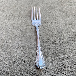 May include: A silver fork with four tines, ornate detailing, and a decorative handle. The fork is made of a shiny, reflective metal and is set against a textured, neutral-toned background. The handle features engraved floral patterns.
