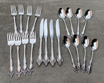 May include: A collection of ornate silver-colored flatware, including forks, knives, and spoons, arranged on a gray surface. The silverware features decorative handles with a floral design, reflecting light. The set includes multiple pieces of each utensil.