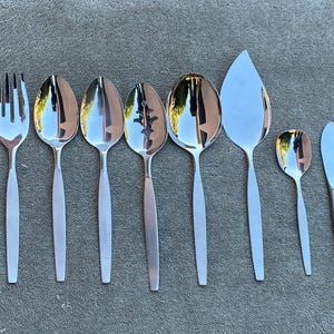 May include: A selection of stainless steel cutlery, including a fork, spoons, and a knife. The cutlery has a simple, modern design with long, slender handles and polished, reflective heads. The set includes serving spoons and a pie server.