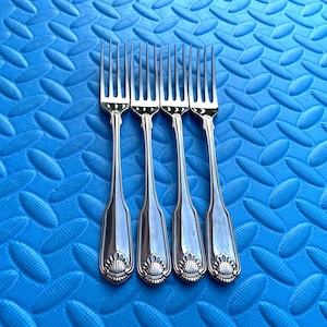Può includere: Quattro forchette d'argento con manici e rebbi decorati, disposte su uno sfondo blu strutturato. Ogni forchetta presenta un motivo a conchiglia alla base del manico. I rebbi sono distanziati uniformemente.