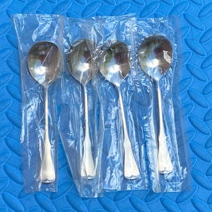 Könnte beinhalten: Vier einzelne silberfarbene Metalllöffel, die jeweils in einer dünnen, durchsichtigen Kunststoffhülle verpackt sind. Die Löffel haben eine klassische abgerundete Laffe und einen glatten, sich verjüngenden Stiel. Das Metall ist poliert.