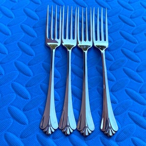May include: Four silver forks with a decorative floral design on the handle are arranged on a textured blue surface. The forks have four tines and a polished finish, reflecting the light.